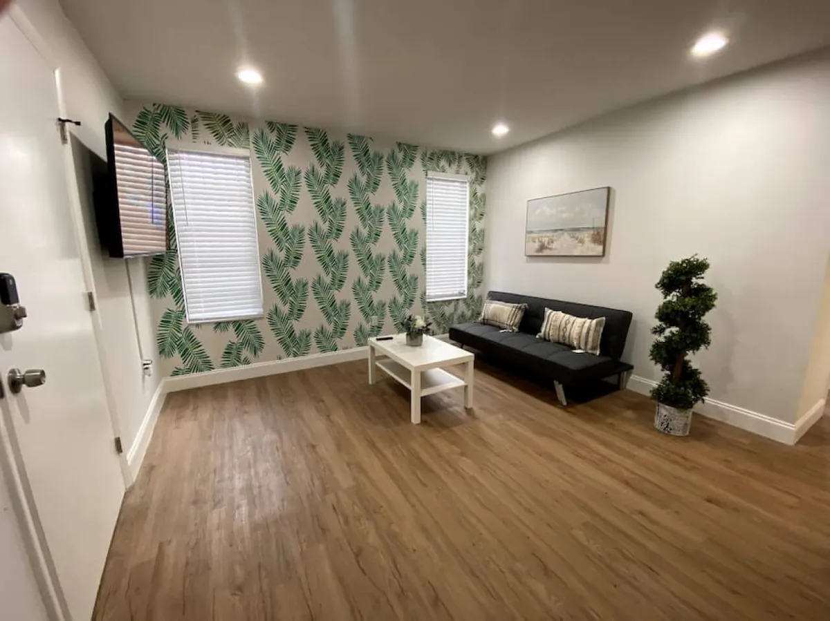 The image features the apartment's inviting living area, anchored by a striking tropical leaf-patterned accent wall. A sleek black futon with striped cushions provides a cozy spot to relax, complemented by a white coffee table with a decorative plant. A wall-mounted flat screen TV offers entertainment, while natural light and modern decor create a fresh, welcoming vibe.