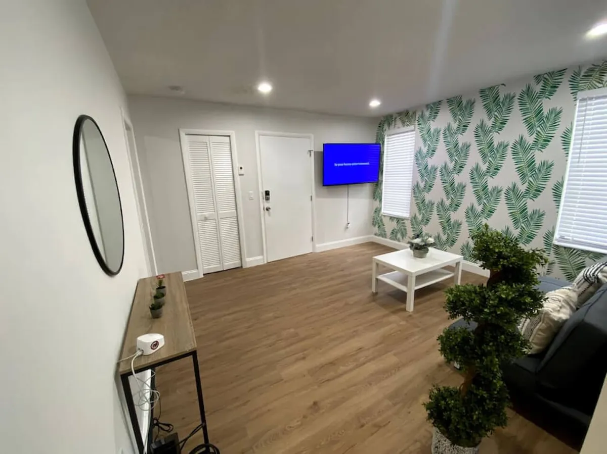 A modern living area featuring a wall-mounted Smart TV, a large decorative mirror, tropical leaf-patterned wallpaper, and a white coffee table. The entry area is visible, suggesting proximity to the entrance and hinting at the presence of a smart lock for seamless check-in.