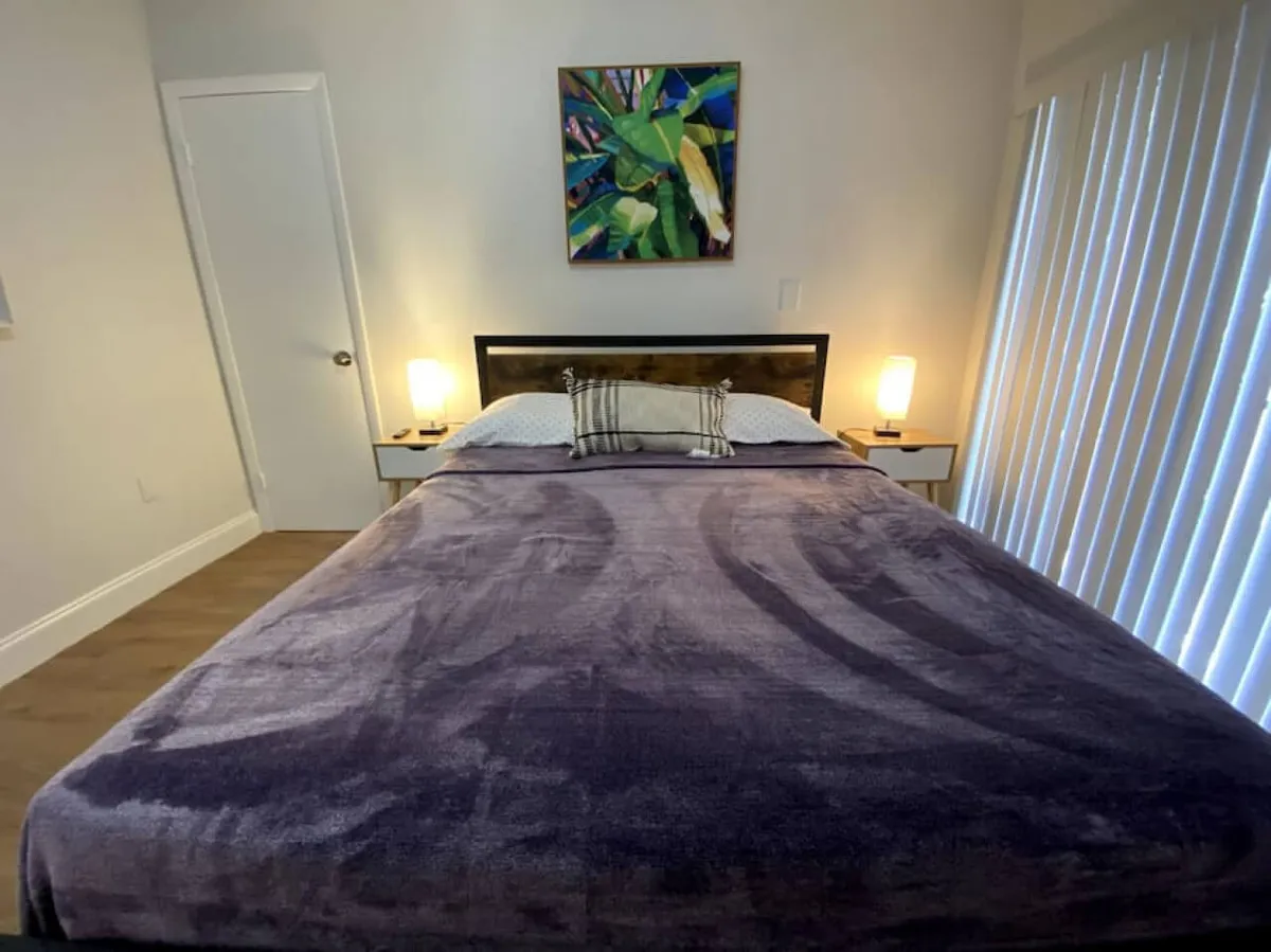 A cozy dining area is visible in the background, adjoining a spacious bedroom with a large bed, tropical-themed artwork, and bedside tables. While the main focus is on the bedroom, the adjoining space hints at a communal area suitable for meals and conversation.
