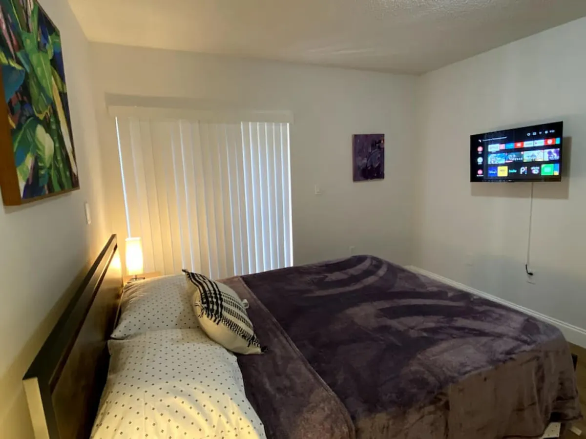A restful bedroom with a king-sized bed adorned with patterned pillows and a dark blanket, wall-mounted smart TV, and vertical blinds covering the window. The modern bedside lamp emits soft lighting, creating a secure and comfortable environment.