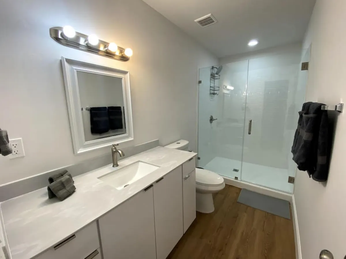 A bright, modern bathroom featuring a large white vanity with a sink and a broad mirror, a glass-enclosed walk-in shower, a toilet, and wood-style flooring. The space is organized, fresh, and inviting, with plenty of room for guests to comfortably use the amenities.