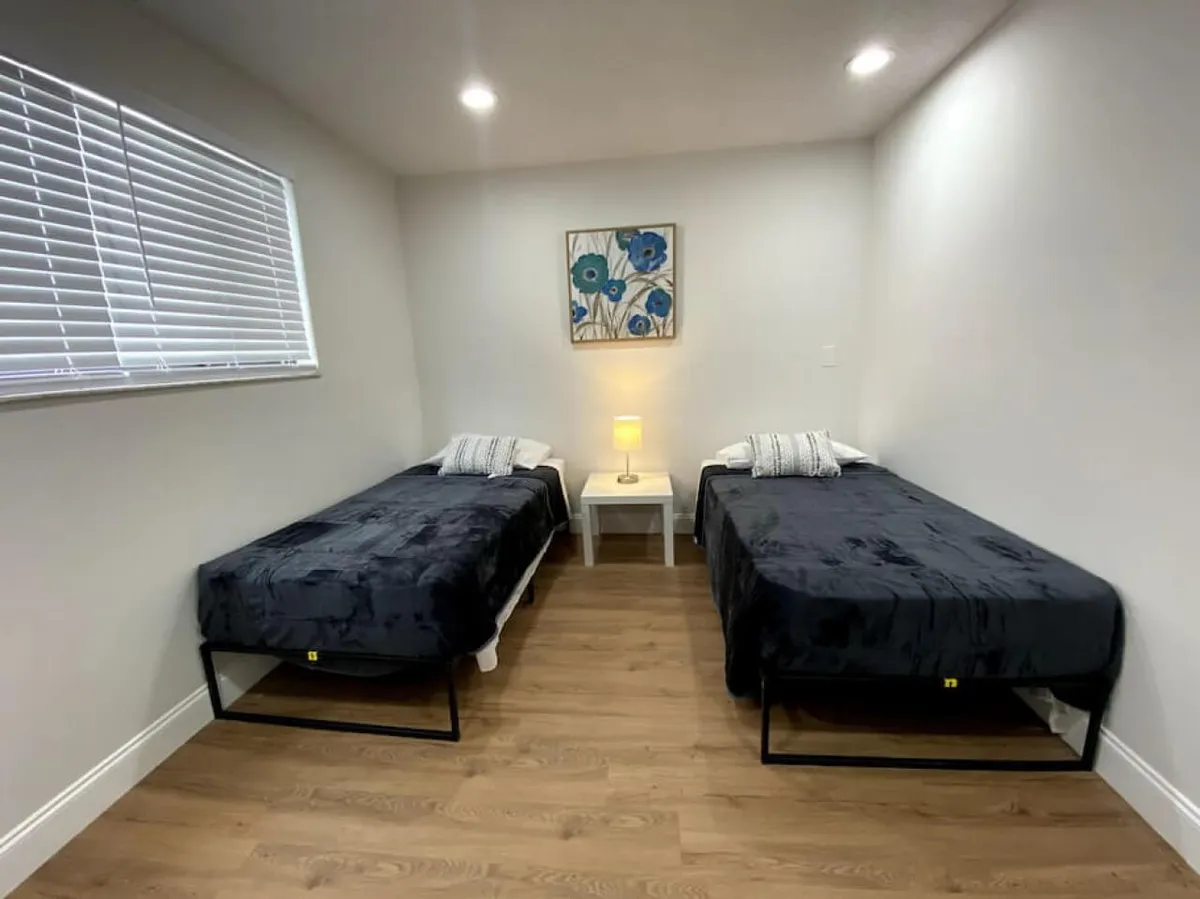 This image shows a serene and minimalistic bedroom featuring two twin beds with sleek black bedding. Between the beds sits a white nightstand topped by a lamp, and blue floral artwork adds a gentle pop of color above. The overall space is uncluttered, bright, and invites relaxation, making it ideal for families, friends, or colleagues sharing the space.