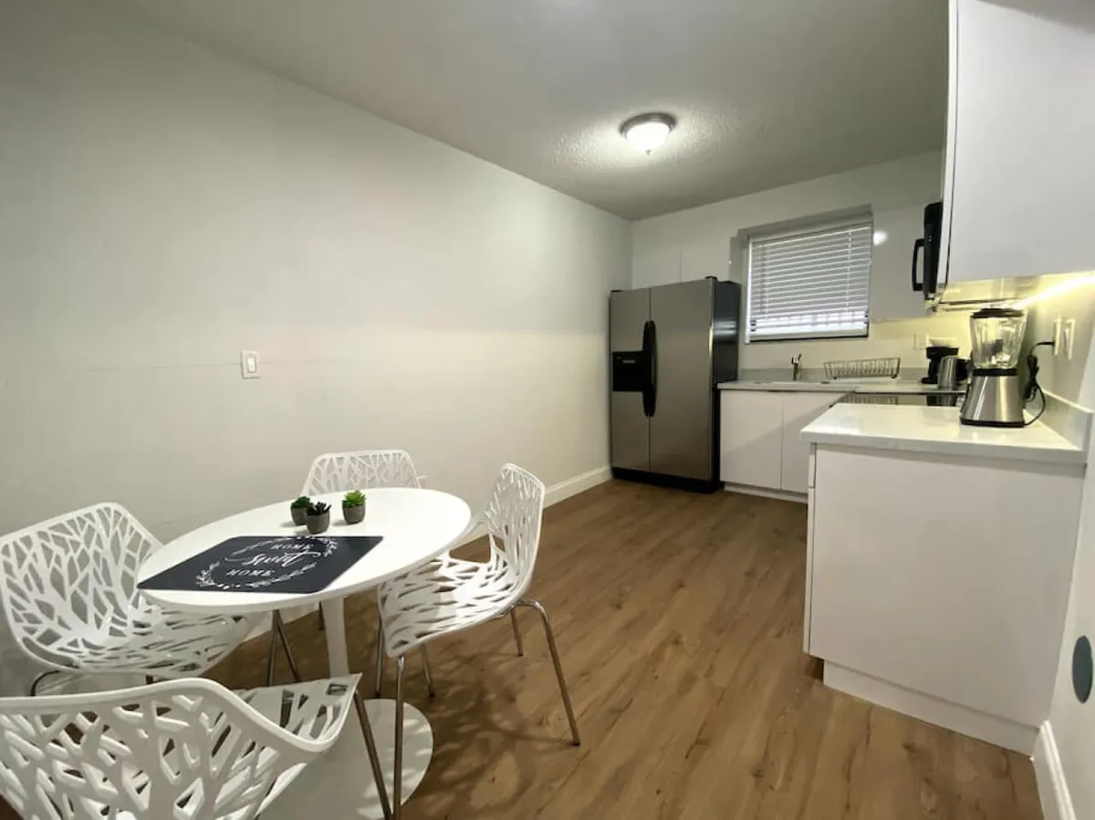 This image showcases a bright, modern kitchen and dining area featuring a round white dining table with uniquely designed white chairs, a stainless steel refrigerator, and sleek white cabinetry. Small appliances like a blender and coffee maker enhance the sense of comfort and convenience. The space feels open and welcoming, ideal for casual dining and socializing.
