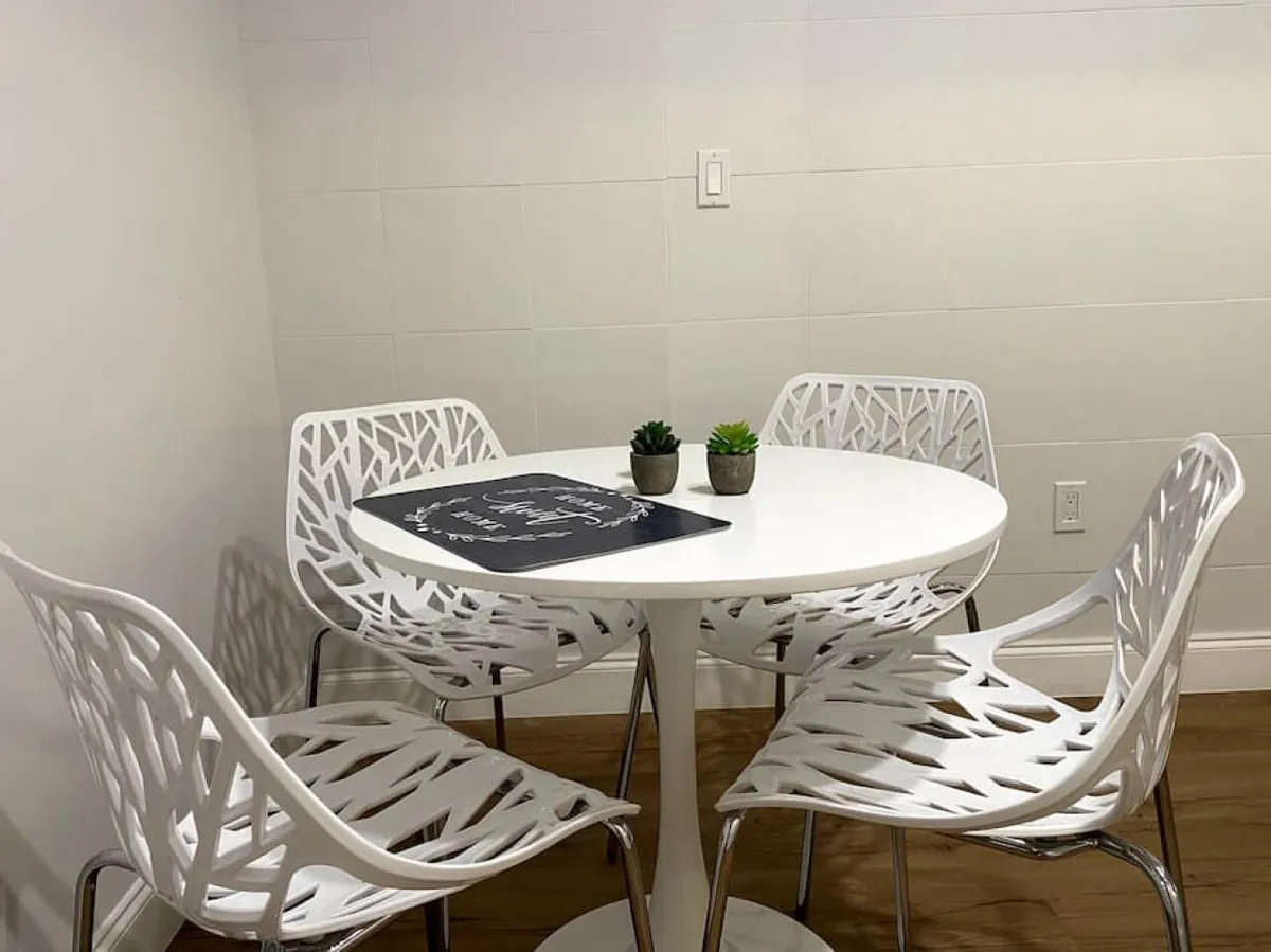 This image features a close-up of a round white dining table surrounded by four modern white chairs with a distinctive cut-out leaf pattern. The table is accessorized with small potted succulents and a black placemat, creating a cozy and inviting spot for informal gatherings, coffee breaks, or planning sessions.